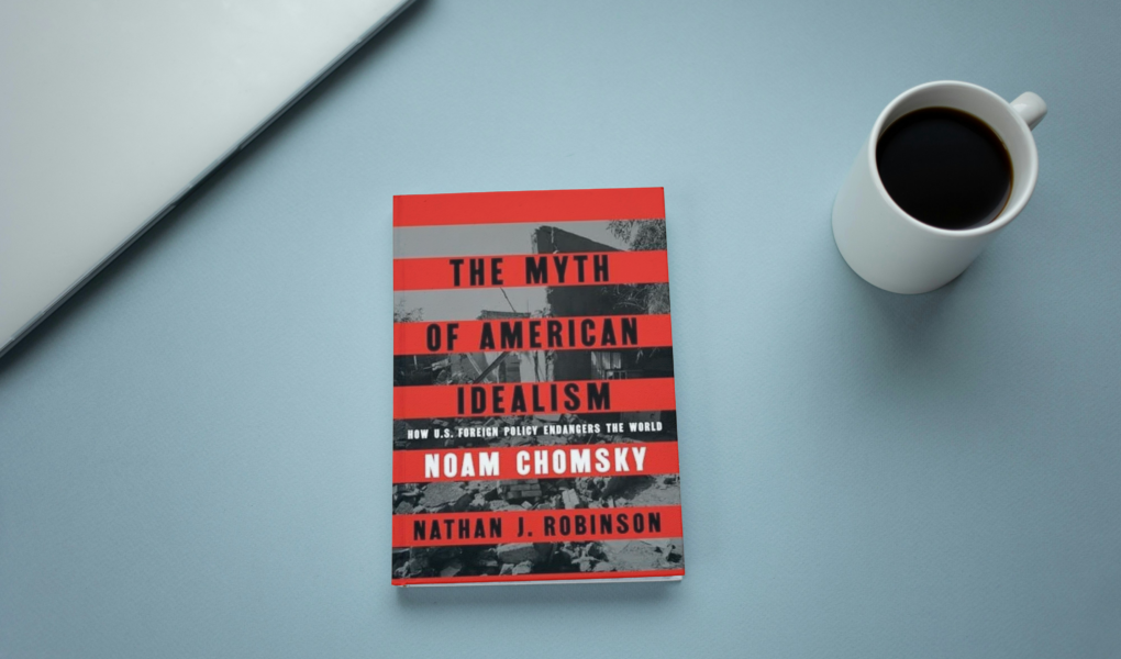 photo of book the myth of American idealism by Noam Chomsky on a blue desk with a laptop and cup of coffee