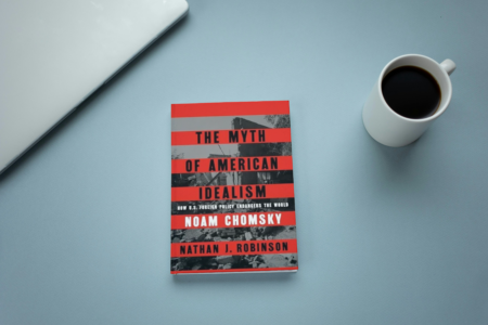 photo of book the myth of American idealism by Noam Chomsky on a blue desk with a laptop and cup of coffee