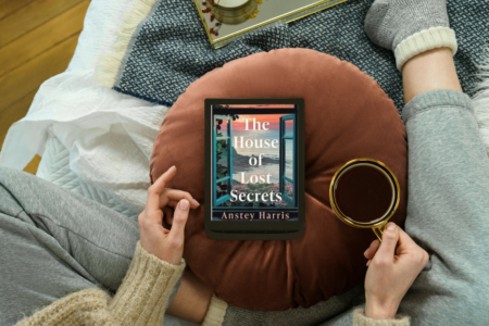 photo of woman sitting on couch with coffee and e-reader with photo of book the house of lost secrets by Anstey harris