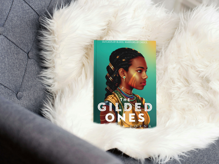 photo of book the gilded ones by namina forna sitting on a white throw blanket in a grey chair